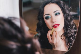 Woman applying red lipstick on lips, looking into mirror. Bright makeup for special occasion