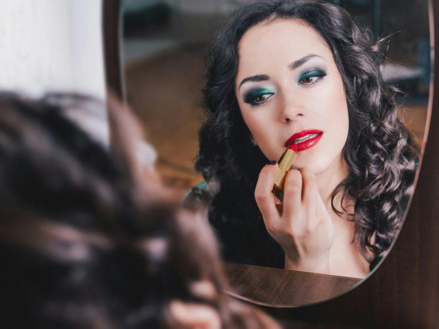 Woman applying red lipstick on lips, looking into mirror. Bright makeup for special occasion