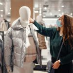 Young brunette lady in face mask chooses warm clothes during pandemic, buying puffy winter jacket